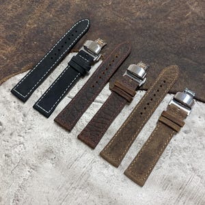 May include: Six leather watch straps in various shades of brown and black are displayed. The straps have stainless steel clasps and are made of textured leather. One black strap has white stitching. The straps are of different widths and textures.