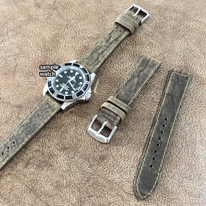 Op de afbeelding: Een bruin leren horlogebandje met een gestructureerd oppervlak. Het bandje is getoond op een voorbeeldhorloge met een zwarte wijzerplaat en zilveren wijzers. Het bandje is ook getoond in twee aparte delen, een met een gesp en een zonder.