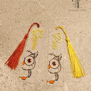 May include: Two clear acrylic bookmarks with gold owl illustrations and gold lettering. The bookmarks have red and yellow tassels.