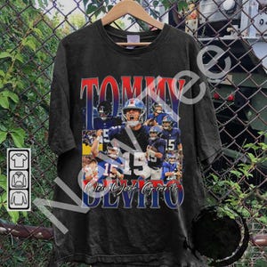 Puede incluir: Camiseta negra con un collage de jugadores de fútbol americano en azul y blanco. La camiseta muestra el nombre "TOMMY" en rojo y "New York Giants" en azul. El número "15" es prominente.