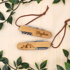 Könnte beinhalten: Zwei Schweizer Taschenmesser aus Holz mit braunen Lederbändern. Ein Messer ist mit dem Namen "Stephan" graviert. Das andere Messer hat ein Bergketten-Design. Beide Messer haben mehrere Werkzeuge und eine helle Holzoberfläche.