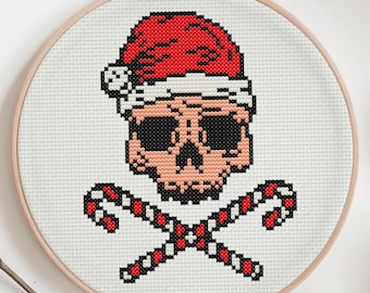May include: A cross-stitch embroidery in a wooden hoop features a skull wearing a Santa hat, with crossed candy canes below. The skull is peach-colored with black eyes and a red hat. The candy canes are red and white. The background is white.