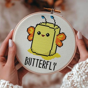 May include: A cross-stitch embroidery hoop with a yellow butterfly design and orange wings, with the word "BUTTERFLY" below. The design is on white fabric, held in a wooden hoop. The background is blurred.