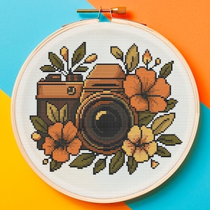 Puede incluir: Un bastidor de bordado con punto de cruz que presenta un diseño de cámara vintage rodeado de flores naranjas y amarillas y hojas verdes. La cámara es marrón y negra, sobre un fondo blanco. El bastidor está sobre un fondo azul y naranja.