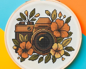 May include: A cross-stitch embroidery hoop featuring a vintage camera design surrounded by orange and yellow flowers and green leaves. The camera is brown and black, and the background is white. The hoop is set against a blue and orange background.