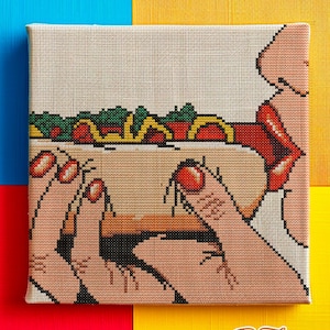 May include: A square canvas cross-stitch artwork depicts a person eating a hot dog. The hot dog has green, red, and yellow toppings. The person's lips are red, and their nails are painted red. The artwork is set against a colorful background.