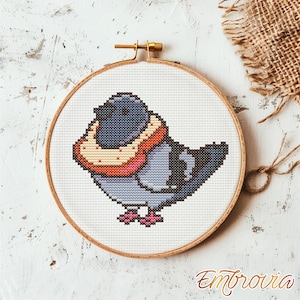 Puede incluir: Bordado de punto de cruz de una paloma de dibujos animados en un aro de madera. El ave es principalmente gris con un collar melocotón y rosa. El bordado está sobre tela blanca y el aro se asegura con un cierre de latón.