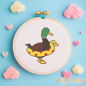 May include: A cross-stitch embroidery hoop showing a duck wearing a yellow inflatable ring with red spots. The duck has a green head and brown body. The background is light blue with yarn clouds and hearts.