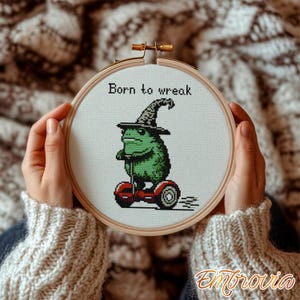 May include: Cross-stitch embroidery in a wooden hoop featuring a green frog wearing a witch hat and riding a red hoverboard. The text "Born to wreak" is above the frog. The embroidery is held by hands wearing a cream-colored sweater.