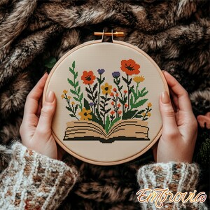 May include: A cross-stitch embroidery hoop featuring a floral design. The design includes an open book with colorful flowers in shades of orange, yellow, purple, and green. The hoop is held by hands, and the text "Embrovia" is visible.