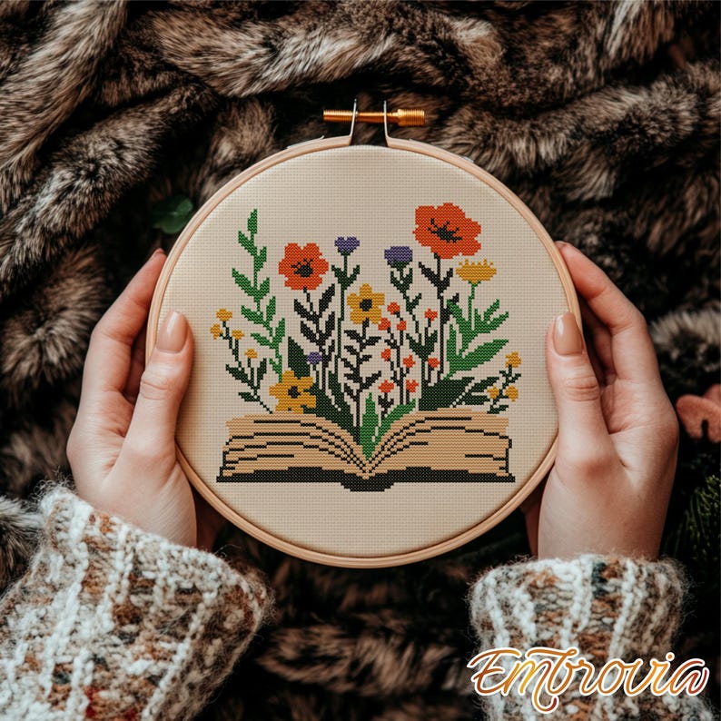 May include: A cross-stitch embroidery hoop featuring a floral design. The design includes an open book with colorful flowers in shades of orange, yellow, purple, and green. The hoop is held by hands, and the text "Embrovia" is visible.