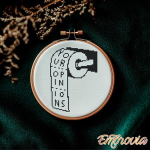 Puede incluir: Un bastidor de bordado con un diseño en blanco y negro de un rollo de papel higiénico. El texto "YOUR OPINIONS" está bordado en el papel. El bastidor tiene un marco de color cobre y está sobre un fondo verde oscuro.