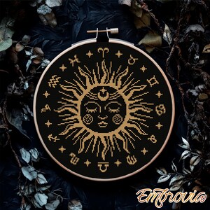 May include: A black cross-stitch embroidery hoop featuring a golden sun design with astrological symbols. The sun has a face and is surrounded by rays and stars. The embroidery is set against a dark background.