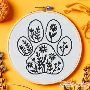 May include: A cross-stitch embroidery hoop with a black paw print design filled with floral patterns. The design is on white fabric, set in a wooden hoop. The background is a bright yellow-orange.