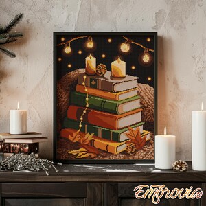 May include: A framed cross-stitch artwork depicts a stack of books with autumn leaves, lit candles, and string lights against a dark background. The artwork has a cozy, warm aesthetic, with a black frame and the word "Embrovia" at the bottom.