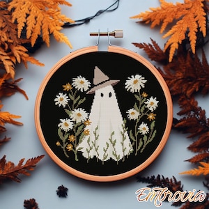 May include: A cross-stitch embroidery featuring a white ghost wearing a witch hat, surrounded by white daisies and green foliage, set against a black background. The design is framed in a wooden hoop, perfect for Halloween decor.