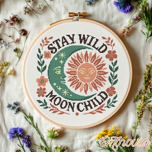 Op de afbeelding: Een borduurring met kruissteek met de tekst "STAY WILD MOON CHILD", een maan, een zon en bloemmotieven in groen, oranje en bruin. De borduursel zit in een houten ring, omringd door bloemen.