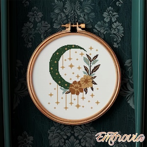 May include: A cross-stitch embroidery in a gold hoop features a crescent moon, stars, and floral elements. The moon is dark green, and the flowers are tan and brown. The embroidery is set against a white background.