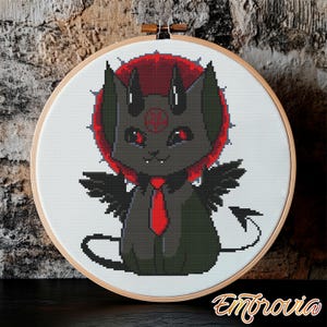 May include: Cross-stitch embroidery in a wooden hoop features a black cat-like creature with red eyes, horns, and wings. The cat wears a red tie and is set against a red halo. The design is on white fabric. The word "Embrovia" is in the bottom right corner.