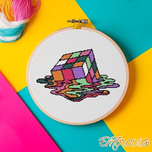 May include: A cross-stitch embroidery hoop featuring a colorful, melting Rubik's Cube design. The cube is rendered in shades of pink, purple, green, orange, and black, with a dripping effect. The background is a vibrant mix of yellow, teal, and pink.