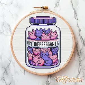 Peut inclure: Broderie au point de croix dans un cercle en bois. Le motif représente un bocal étiqueté "ANTIDÉPRESSEURS" rempli de chats de dessin animé roses et violets. Le bocal a un couvercle violet et repose sur un fond en tissu blanc.