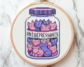 May include: Cross-stitch art in a wooden hoop. The design features a jar labeled "ANTIDEPRESSANTS" filled with pink and purple cartoon cats. The jar has a purple lid and sits on a white fabric background.
