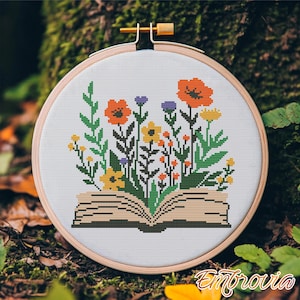 Op de afbeelding: Een kruissteek borduurring met een bloemenontwerp. Het ontwerp toont een open boek met bloemen en groen. De bloemen zijn oranje, geel en paars. De ring is gemaakt van hout en heeft een goudkleurige stelschroef.