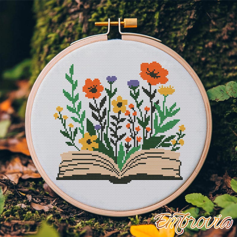 May include: A cross-stitch embroidery hoop featuring a colorful floral design. The design depicts an open book with flowers and greenery sprouting from it. The flowers are orange, yellow, and purple. The hoop is made of wood and has a gold-colored adjustment screw.