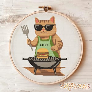 May include: Cross-stitch embroidery of a cat wearing sunglasses and a green apron that says "CHEF". The cat is holding a fork and grilling a hamburger on a small barbecue grill. The embroidery is set in a wooden hoop.