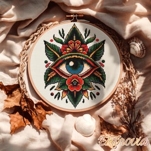 May include: A cross-stitch embroidery of an eye with a blue iris, framed by green leaves and red flowers. The design is set in a wooden hoop, with a rope border. The word "Embrovia" is in the bottom right corner.