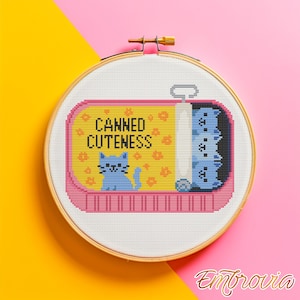 May include: A cross-stitch embroidery hoop with a playful design. The artwork shows a pink and yellow sardine can with the text "Canned Cuteness" and a blue cat illustration. Inside the can are stacked blue cat figures. The background is a gradient of yellow and pink.