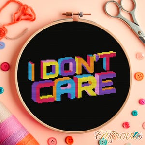 May include: A black cross-stitch hoop with the phrase "I DON'T CARE" in colorful, 3D-style lettering. The embroidery is set against a black background, surrounded by a wooden hoop. The image includes scissors, thread, and buttons.