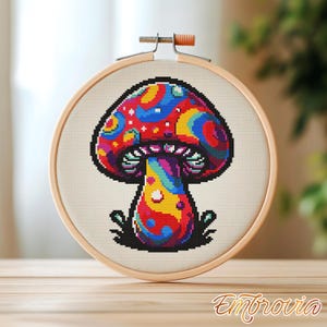 May include: A colorful cross-stitch mushroom design in a wooden embroidery hoop. The mushroom features a vibrant pattern of red, blue, yellow, and purple. The embroidery hoop is made of light-colored wood, and the fabric is a neutral beige.