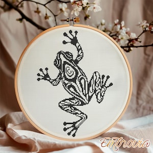 May include: A black and white cross-stitch frog design in a wooden embroidery hoop. The frog has an intricate pattern. The background is a neutral color with a branch of white flowers. The word "Embrovia" is in the bottom right corner.