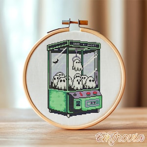 May include: Cross-stitch embroidery of a claw machine with a ghost theme. The machine is green with the text "Grab a Ghost". Several white ghosts are inside. The embroidery is set in a wooden hoop.