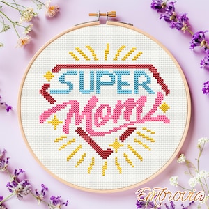 May include: A cross-stitch embroidery hoop with the words "SUPER Mom" in blue and pink, surrounded by yellow and red accents. The design is set against a white fabric background, framed by a wooden hoop.