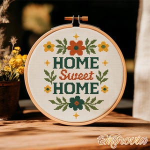May include: A cross-stitch embroidery hoop with the words "HOME Sweet HOME" in green and orange. The design includes floral and leaf motifs in yellow, orange, and green. The embroidery is set in a wooden hoop, perfect for home decor.