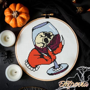 Könnte beinhalten: Kreuzstich-Stickrahmen mit einem Totenkopf, der ein Weinglas hält. Der Totenkopf trägt ein rotes Outfit und einen lila Schal. Der Hintergrund ist weiß, umgeben von Halloween-Dekorationen, darunter ein Kürbis und Kerzen.