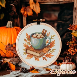 May include: Cross-stitch embroidery in a wooden hoop featuring a teal mug of hot chocolate with marshmallows and cinnamon sticks, resting on a stack of books and surrounded by fall leaves. A pumpkin and other fall decor are in the background.