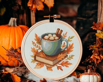May include: Cross-stitch embroidery in a wooden hoop featuring a teal mug of hot chocolate with marshmallows and cinnamon sticks, resting on a stack of books and surrounded by fall leaves. A pumpkin and other fall decor are in the background.