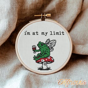May include: Cross-stitch embroidery in a wooden hoop. The design features a green frog with fairy wings, holding a glass of wine, sitting on a red mushroom. The text "i'm at my limit" is above the frog. The embroidery is set against a neutral fabric.
