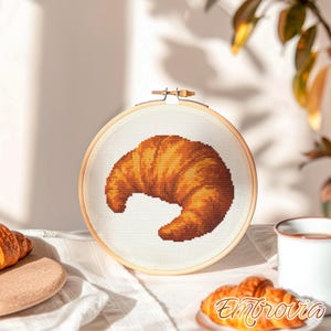 May include: A cross-stitch embroidery hoop featuring a detailed, golden-brown croissant design. The artwork is set against a white fabric background, framed by a natural wood hoop. The image also includes a croissant, a cup, and the text "Embrovia".