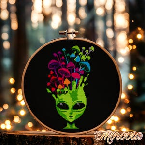 Könnte beinhalten: Kreuzstich-Stickerei mit einem grünen Alien-Kopf mit bunten Pilzen, die aus seinem Kopf sprießen. Das Design befindet sich vor einem schwarzen Hintergrund in einem hölzernen Stickrahmen. Das Kunstwerk wird auf einer Holzoberfläche präsentiert.