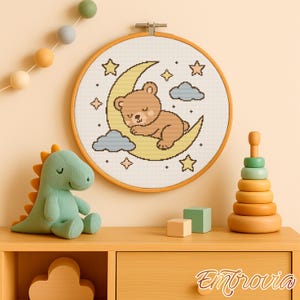 Pode incluir: Um bastidor de bordado em ponto cruz com um urso castanho a dormir numa lua crescente amarela, rodeado por estrelas e nuvens. Um brinquedo de pelúcia de dinossauro verde e um brinquedo de empilhar de madeira estão numa superfície de madeira.