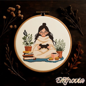 Puede incluir: Bordado de punto de cruz en un aro de madera que representa a una persona con gafas y una manta, sosteniendo un mando de videojuegos. La escena incluye plantas, libros, una taza y galletas. El diseño está sobre un fondo crema.