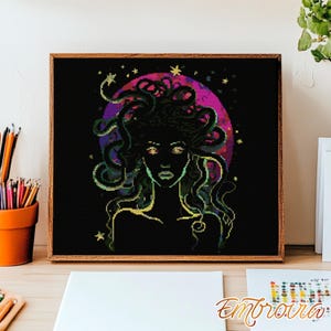 May include: Cross-stitch artwork in a wooden frame, depicting Medusa. The design features a woman with snake hair against a black backdrop with a purple and pink moon. The artwork includes stars and the word "Embroira" in the bottom right corner. The artwork is in a wooden frame.