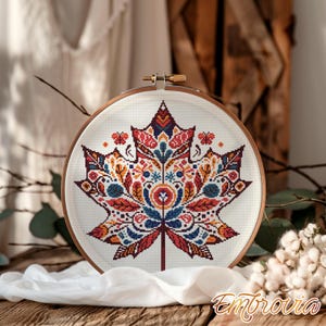 May include: A cross-stitch embroidery of a maple leaf in a wooden hoop. The leaf design features a symmetrical pattern with red, orange, blue, and white details. The embroidery is set against a white fabric background.