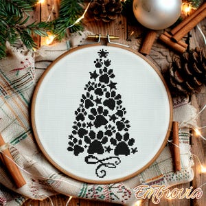 Op de afbeelding: Een borduurring met kruissteek met een kerstboomontwerp gemaakt van zwarte pootafdrukken. De borduursel staat tegen een witte achtergrond en wordt omlijst door een houten ring. Het ontwerp bevat een strik aan de basis en sterren aan de top.