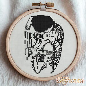 May include: A black and white cross-stitch embroidery of a couple embracing, set in a wooden hoop. The design features intricate details and is mounted on white fabric. The embroidery is a handmade craft.