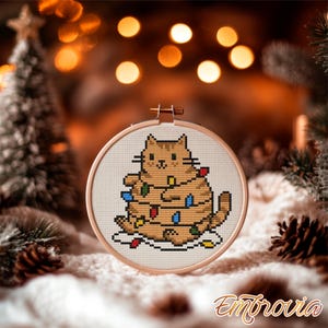 Puede incluir: Un bastidor de bordado de punto de cruz con un gato de dibujos animados adornado con luces navideñas de colores. El gato es marrón con detalles negros, y las luces son rojas, azules y amarillas. Fondo borroso con decoraciones navideñas.